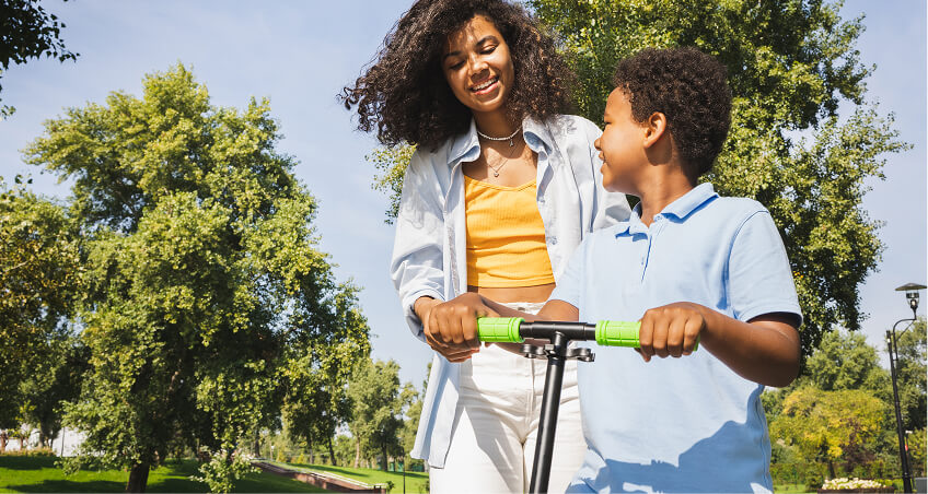 Uma criança com um patinete sendo ajudada pela sua mãe com ambos sorrindo
